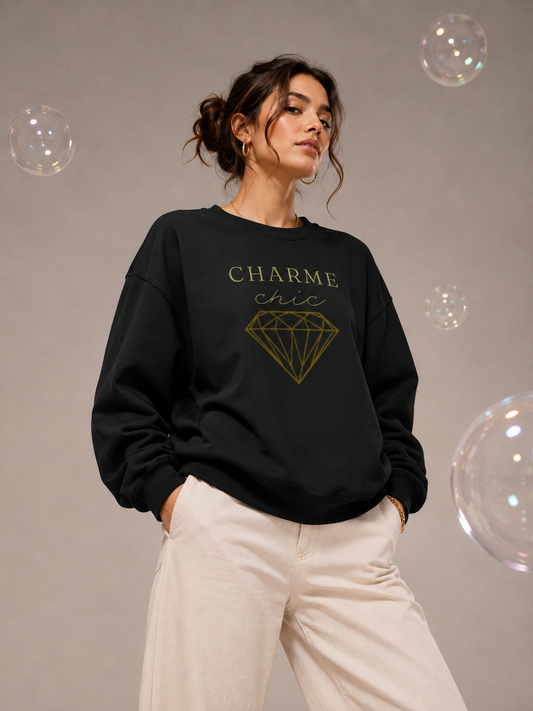 A woman wearing a black Charme Chic graphic sweatshirt with gold typography and a diamond design, paired with cream trousers in a neutral studio setting with floating iridescent bubbles.