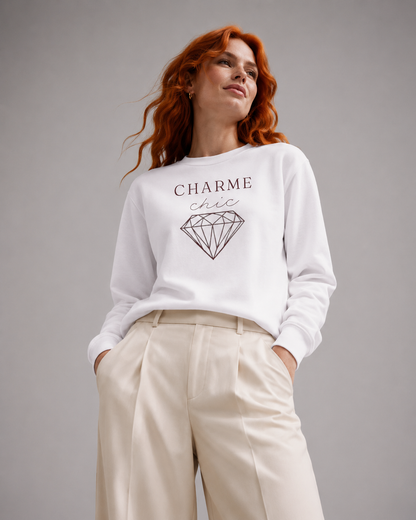 A woman with red hair wearing a white long-sleeve sweatshirt with a black graphic of a diamond and the text 'CHARME chic', paired with cream-colored wide-leg trousers, against a solid gray studio background.
