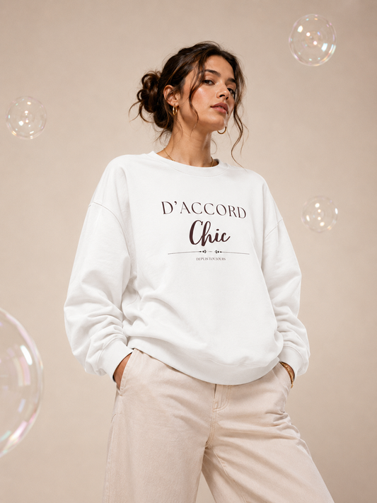A woman wearing a white D’Accord Chic crewneck sweatshirt with elegant chest typography, paired with beige trousers in a warm champagne-beige studio setting with floating iridescent bubbles.