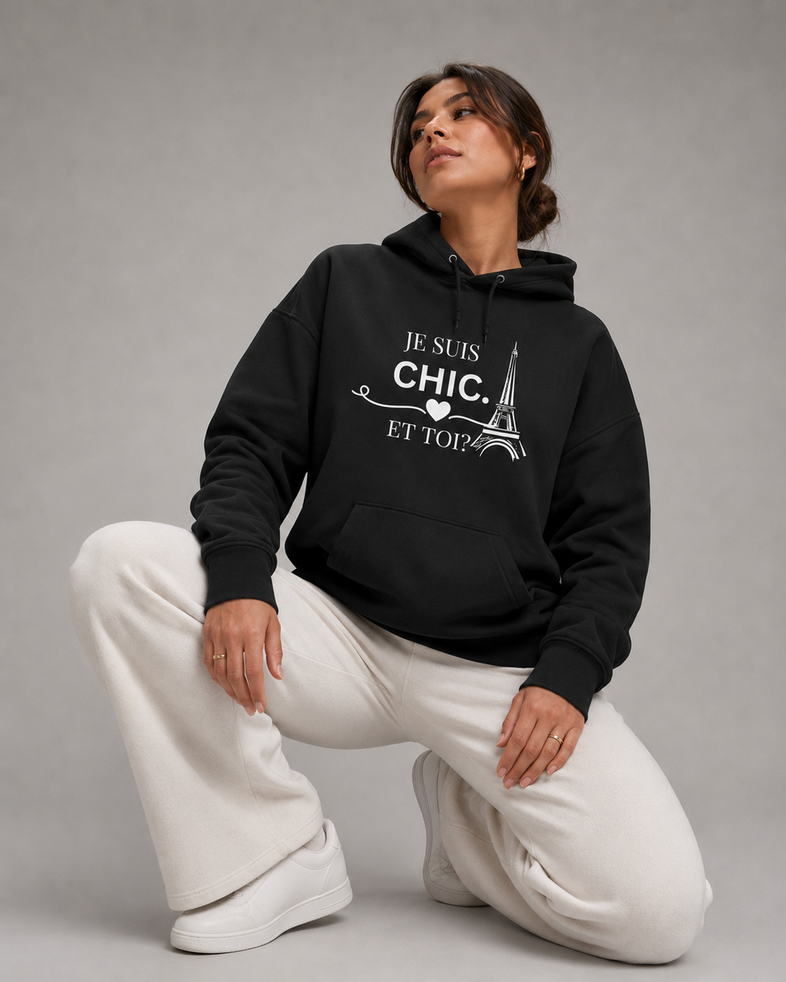 A woman kneels on a gray studio floor wearing a black pullover hoodie with a white graphic print that reads 'JE SUIS CHIC. ET TOP.