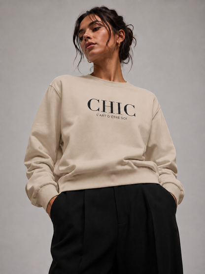 A woman wearing a beige crewneck sweatshirt with “CHIC L’ART D’ÊTRE SOI” printed in black across the chest, paired with black trousers, against a neutral gray studio background.