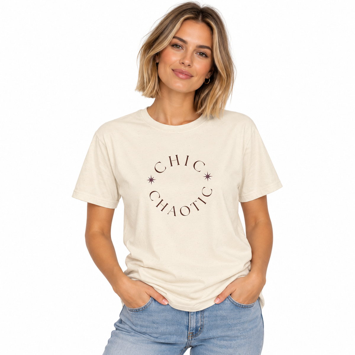 A woman wearing a cream-colored t-shirt with a dark brown graphic that reads 'CHIC CHAOTIC' in a circular arrangement with small stars, paired with light blue jeans, against a plain white background.