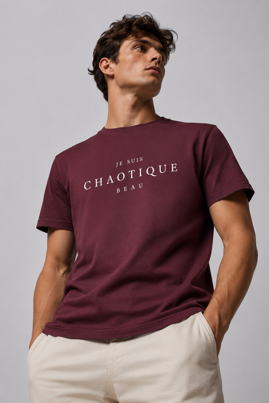 A man wearing a maroon short-sleeve t-shirt with white text reading 'JE SUIS CHAOTIQUE BEAU' on the chest, paired with cream-colored pants, against a plain gray studio background.