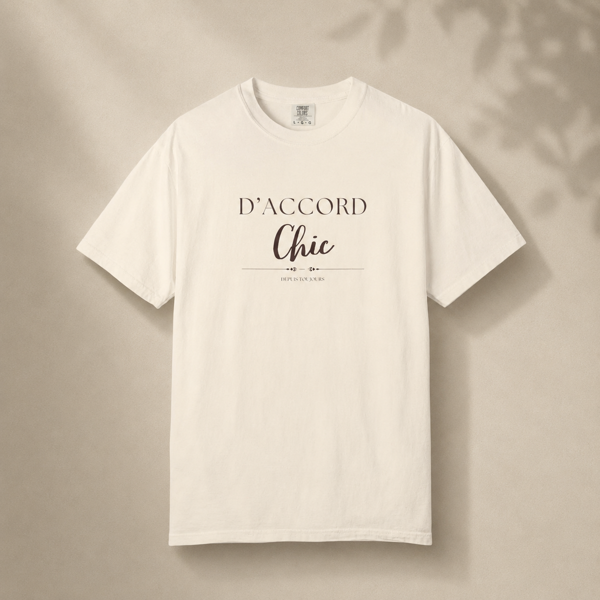 Beige short-sleeve crewneck T-shirt featuring the “D’Accord Chic” graphic on the front, displayed against a warm beige background with soft leaf shadows.