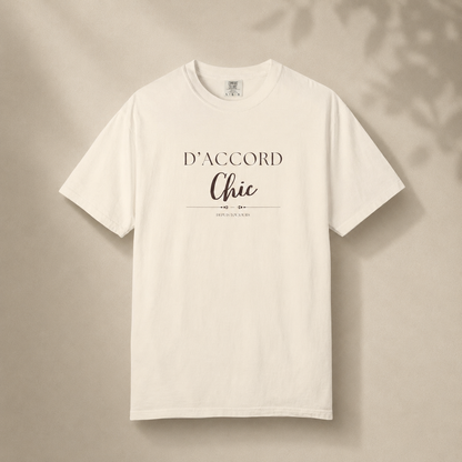 Beige short-sleeve crewneck T-shirt featuring the “D’Accord Chic” graphic on the front, displayed against a warm beige background with soft leaf shadows.