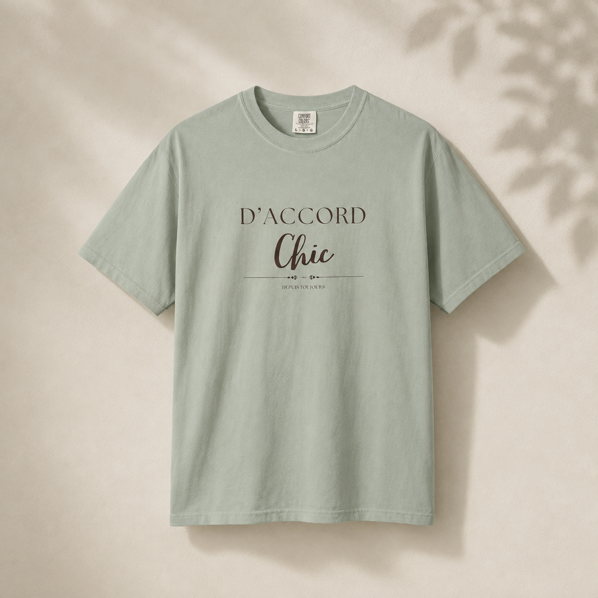 Light green short-sleeve crewneck T-shirt featuring the “D’Accord Chic” graphic on the front, displayed against a warm beige background with soft leaf shadows.