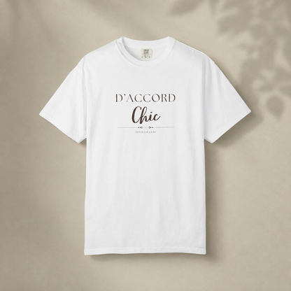 White short-sleeve crewneck T-shirt featuring the “D’Accord Chic” graphic on the front, displayed against a warm beige background with soft leaf shadows.