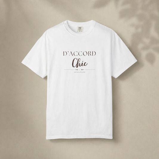 White short-sleeve crewneck T-shirt featuring the “D’Accord Chic” graphic on the front, displayed against a warm beige background with soft leaf shadows.