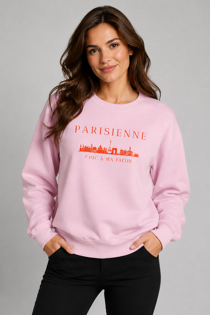 A woman wearing a light pink crewneck sweatshirt with a bright orange “PARISIENNE” graphic, Paris skyline design, and “CHIC À MA FAÇON” text, paired with light-wash blue jeans against a neutral gray studio background.