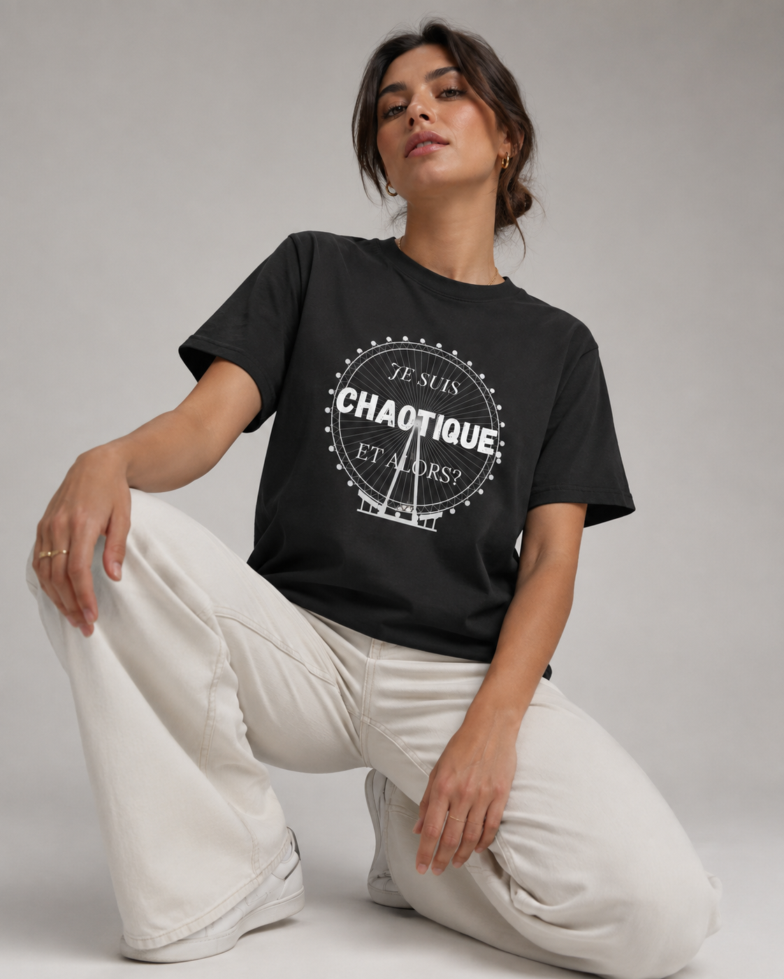 A woman kneels on a light gray studio floor wearing a black graphic T-shirt and off-white wide-leg pants. The shirt has a white circular graphic with the words 'JE SUIS CHAOTIQUE ET ALORSI'.