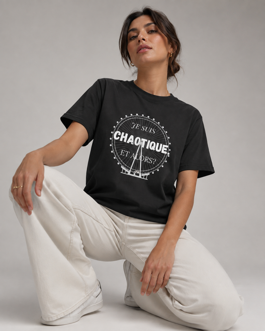 A woman kneels on a light gray studio floor wearing a black graphic T-shirt and off-white wide-leg pants. The shirt has a white circular graphic with the words 'JE SUIS CHAOTIQUE ET ALORSI'.