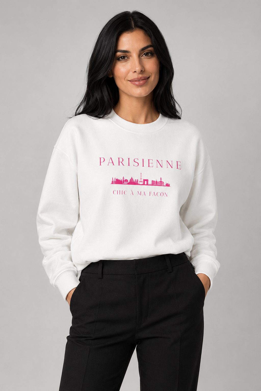 A woman wearing a off-white crewneck sweatshirt with a bright pink “PARISIENNE” graphic, Paris skyline design, and “CHIC À MA FAÇON” text, paired with light-wash blue jeans against a neutral gray studio background.