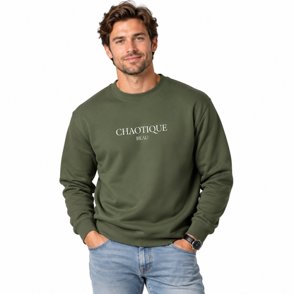A man wearing an olive green crewneck sweatshirt with 'CHAOTIQUE BEAU' printed in white on the chest, paired with light blue jeans, against a plain white background.