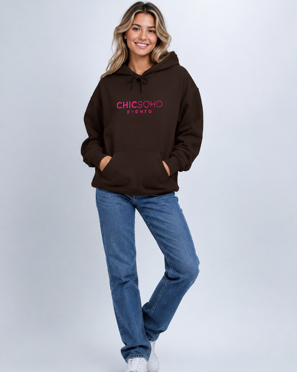 A woman modeling a dark brown pullover hoodie with 'CHICSOHO BYCHTQ' printed in pink and white on the chest, paired with blue jeans and white sneakers, against a light gray studio background.