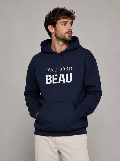 A man wearing a navy blue hoodie with 'D'ACCORD BEAU' printed in white on the chest, paired with off-white trousers, against a plain gray studio background.