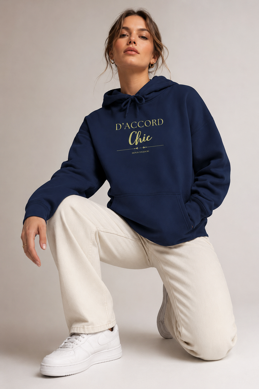 A woman wearing a navy blue D’Accord Chic hoodie with gold chest typography, cream trousers and white sneakers, kneeling on a light grey studio floor.