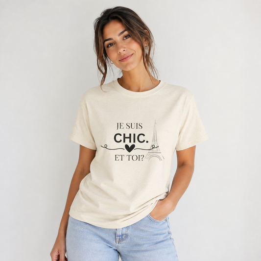 A woman wearing a cream “Je Suis Chic. Et Toi?” graphic T-shirt with black text and an Eiffel Tower illustration, paired with light blue jeans against a light gray studio background.