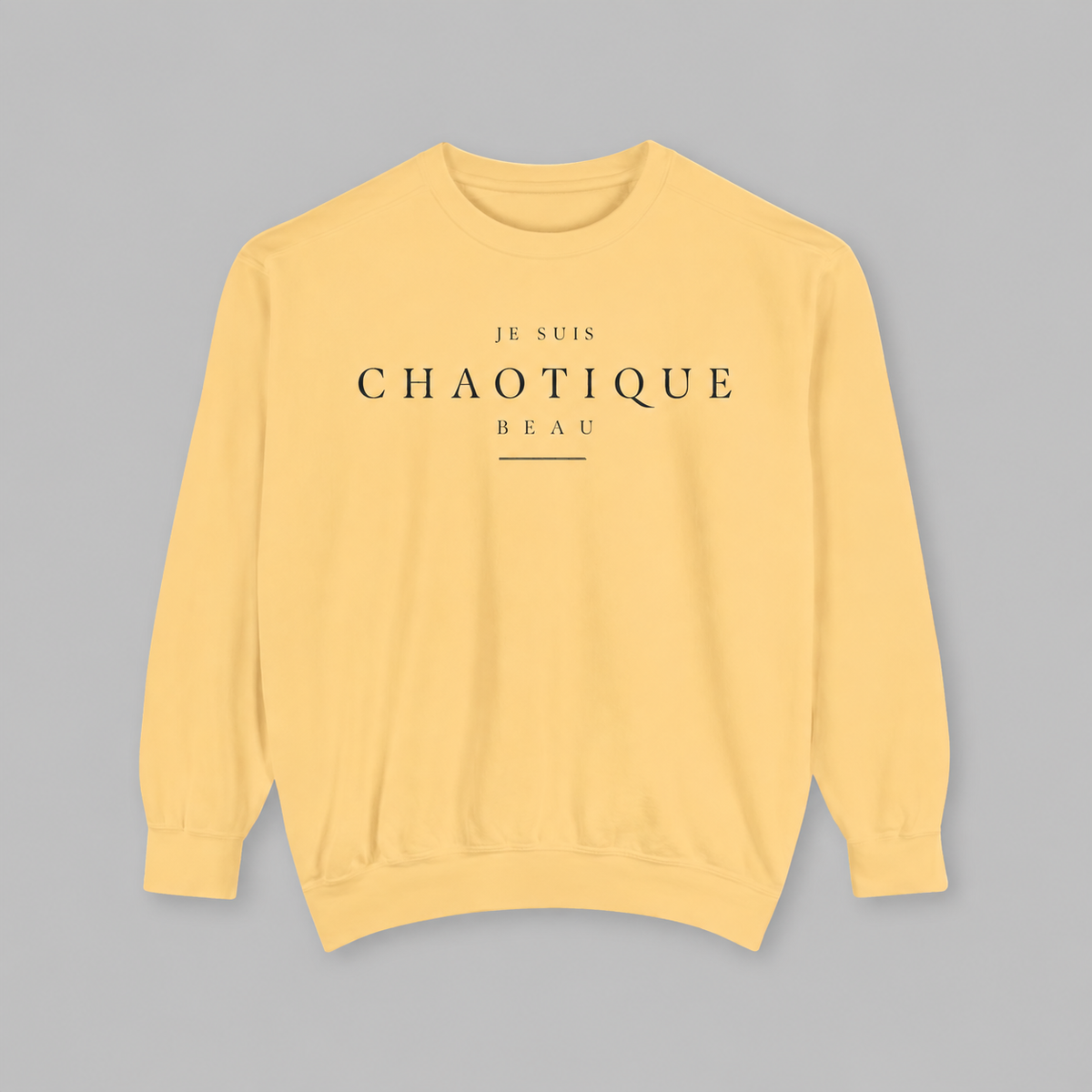 A pale yellow crewneck sweatshirt with the French text 'JE SUIS CHAOTIQUE BEAU' printed in black on the chest, displayed flat against a solid gray background.