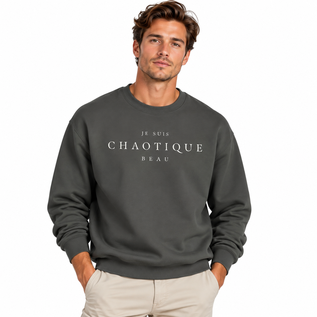 A man wearing a dark gray crewneck sweatshirt with white text reading 'JE SUIS CHAOTIQUE BEAU' on the chest, paired with light beige pants, against a plain white background.