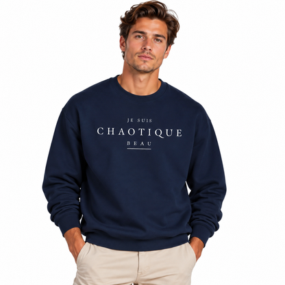 A man wearing a navy blue crewneck sweatshirt with white printed text reading 'JE SUIS CHAOTIQUE BEAU' on the chest, paired with light beige pants, against a plain white background.