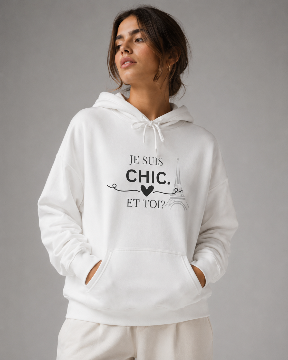 A woman wearing a white pullover hoodie with a black graphic print that reads 'JE SUIS CHIC. ET TOI?' with a small heart and decorative swirls, posed against a neutral gray studio background.