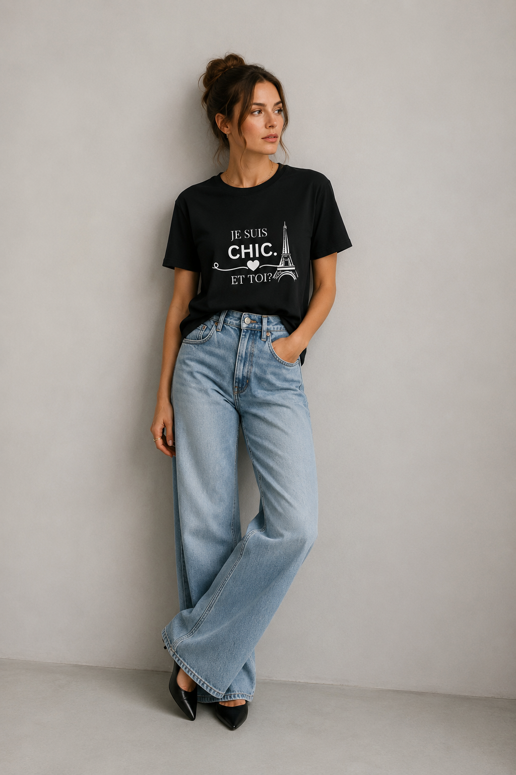Woman wearing a black “Je Suis Chic. Et Toi?” graphic T-shirt with wide-leg light-wash jeans in a minimalist light gray studio.