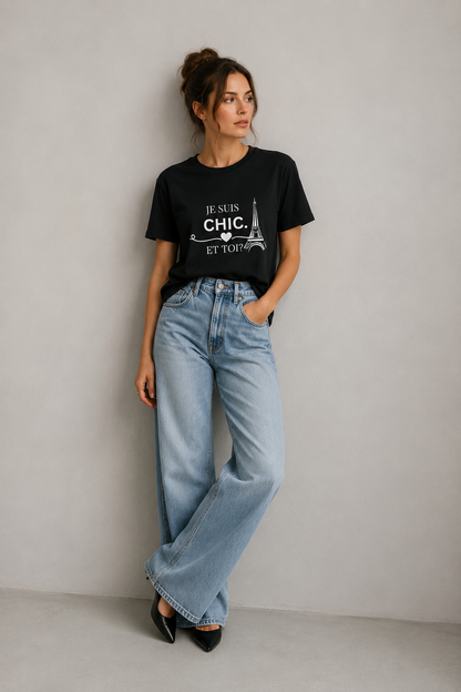 Woman wearing a black “Je Suis Chic. Et Toi?” graphic T-shirt with wide-leg light-wash jeans in a minimalist light gray studio.