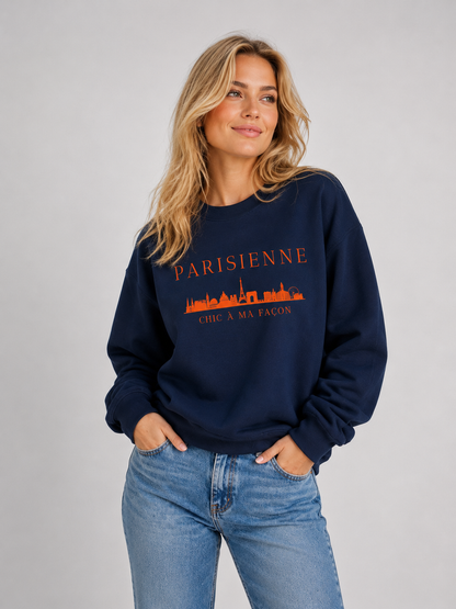 Woman wearing the PARISIENNE Chic à Ma Façon Paris Graphic Sweatshirt in a navy shade with a bright orange Paris skyline print, styled with light blue jeans in a light studio setting.