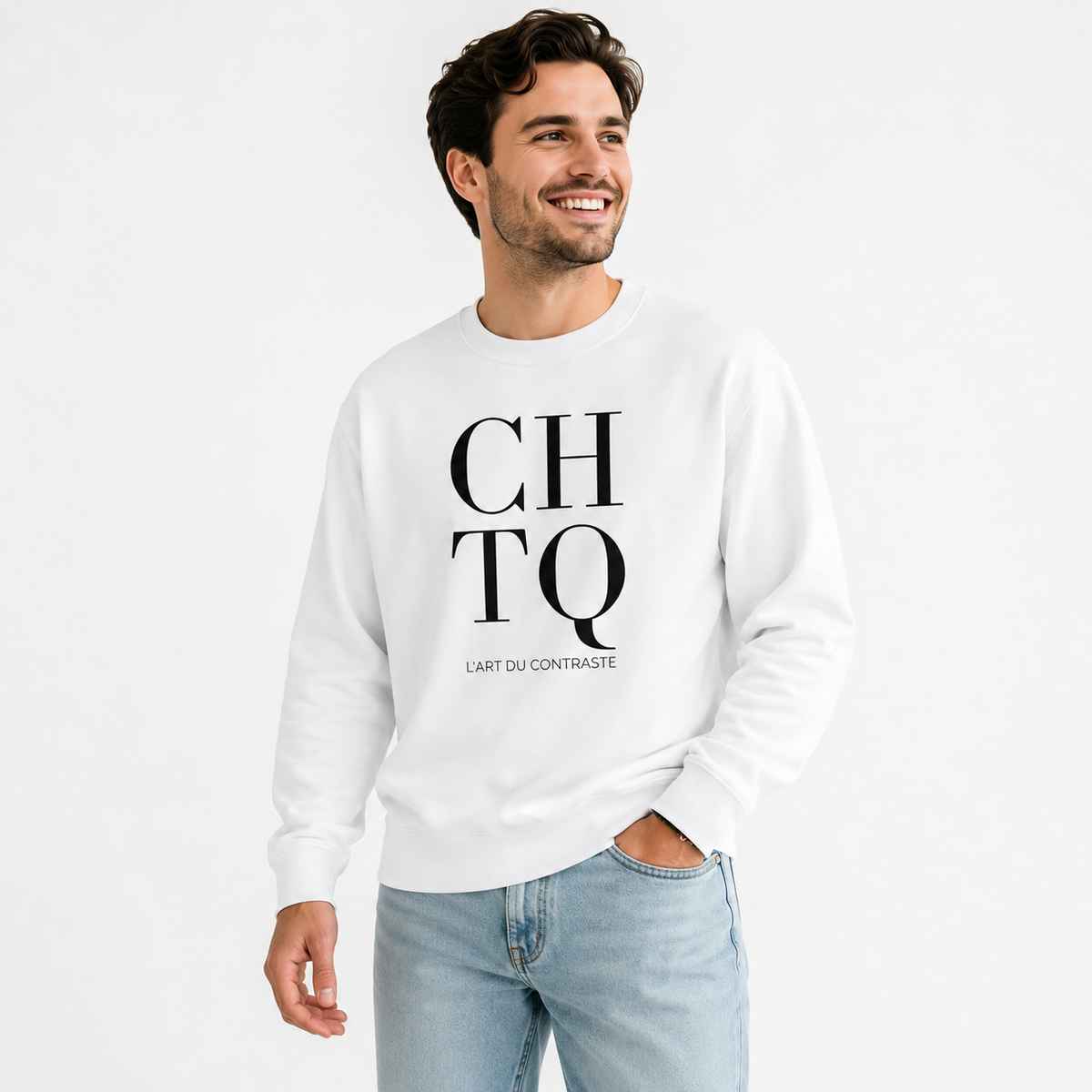 A smiling man wearing a white CHTQ graphic crewneck sweatshirt with black “CHTQ” and “L’ART DU CONTRASTE” print, paired with light blue jeans on a clean white studio background.