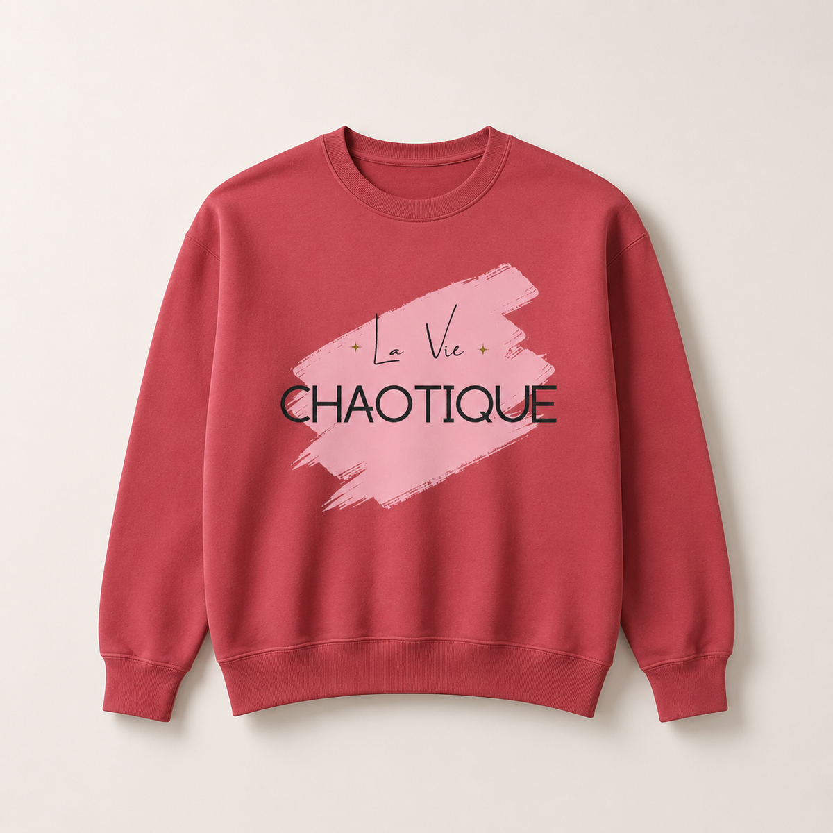 Dusty rose crewneck sweatshirt featuring the “La Vie Chaotique” graphic in black over a soft pink brushstroke on the front, displayed against a light beige background.