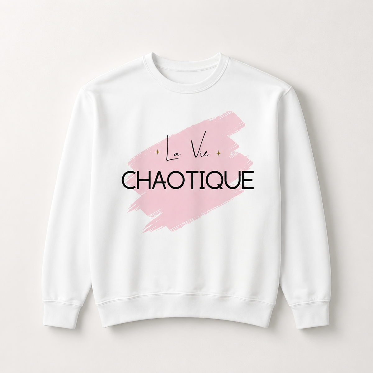 White crewneck sweatshirt featuring the “La Vie Chaotique” graphic in black over a soft pink brushstroke on the front, displayed against a light beige background.