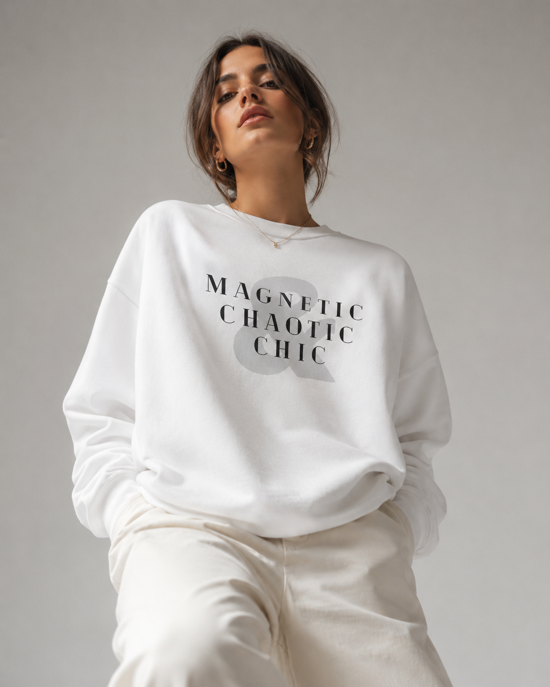 A woman wearing a white Magnetic Chaotic Chic crewneck sweatshirt with bold black graphic typography, paired with white trousers against a neutral grey studio background.