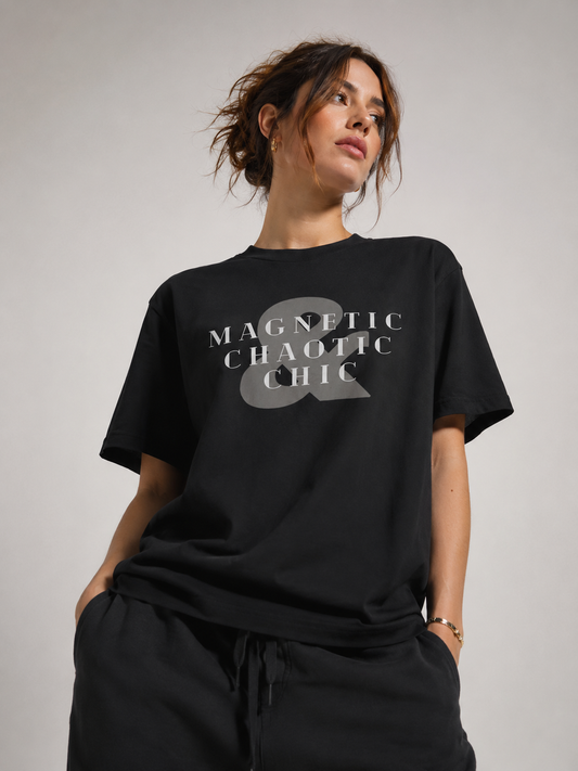 Woman wearing an oversized black graphic T-shirt featuring the text “Magnetic Chaotic Chic” and a large gray ampersand, paired with black drawstring pants in a light gray studio.