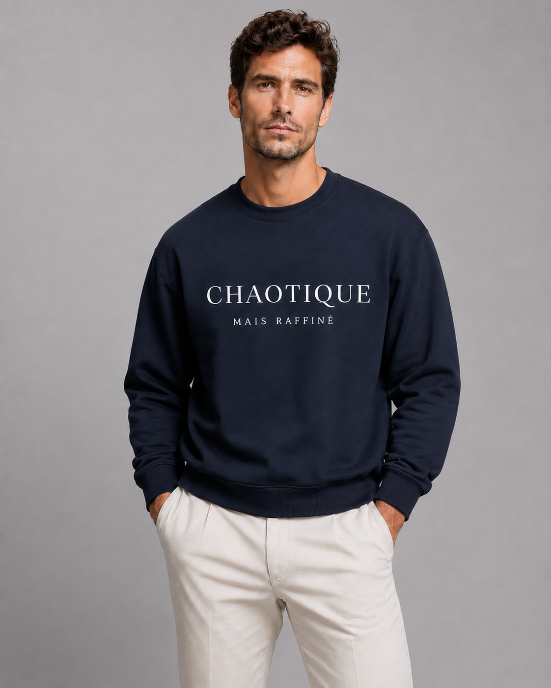 A man wearing a navy blue crewneck sweatshirt with 'CHAOTIQUE MAIS RAFFINE' printed in white across the chest, paired with off-white trousers, against a solid gray studio background.
