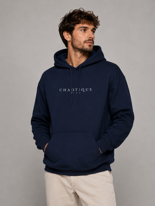 A man wearing a navy blue hoodie with 'CHAOTIQUE BEAU' printed in white on the chest, paired with light beige pants, set against a plain gray studio background.