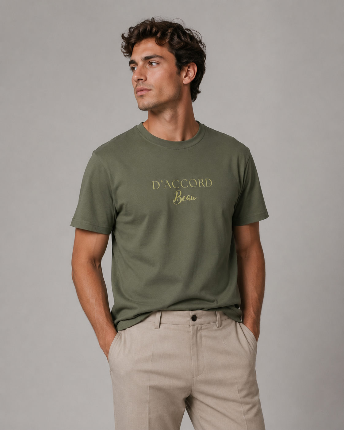 A man wearing an olive green crewneck t-shirt with gold foil text reading 'D'ACCORD Beau' on the chest, paired with beige trousers, photographed against a neutral gray studio background.