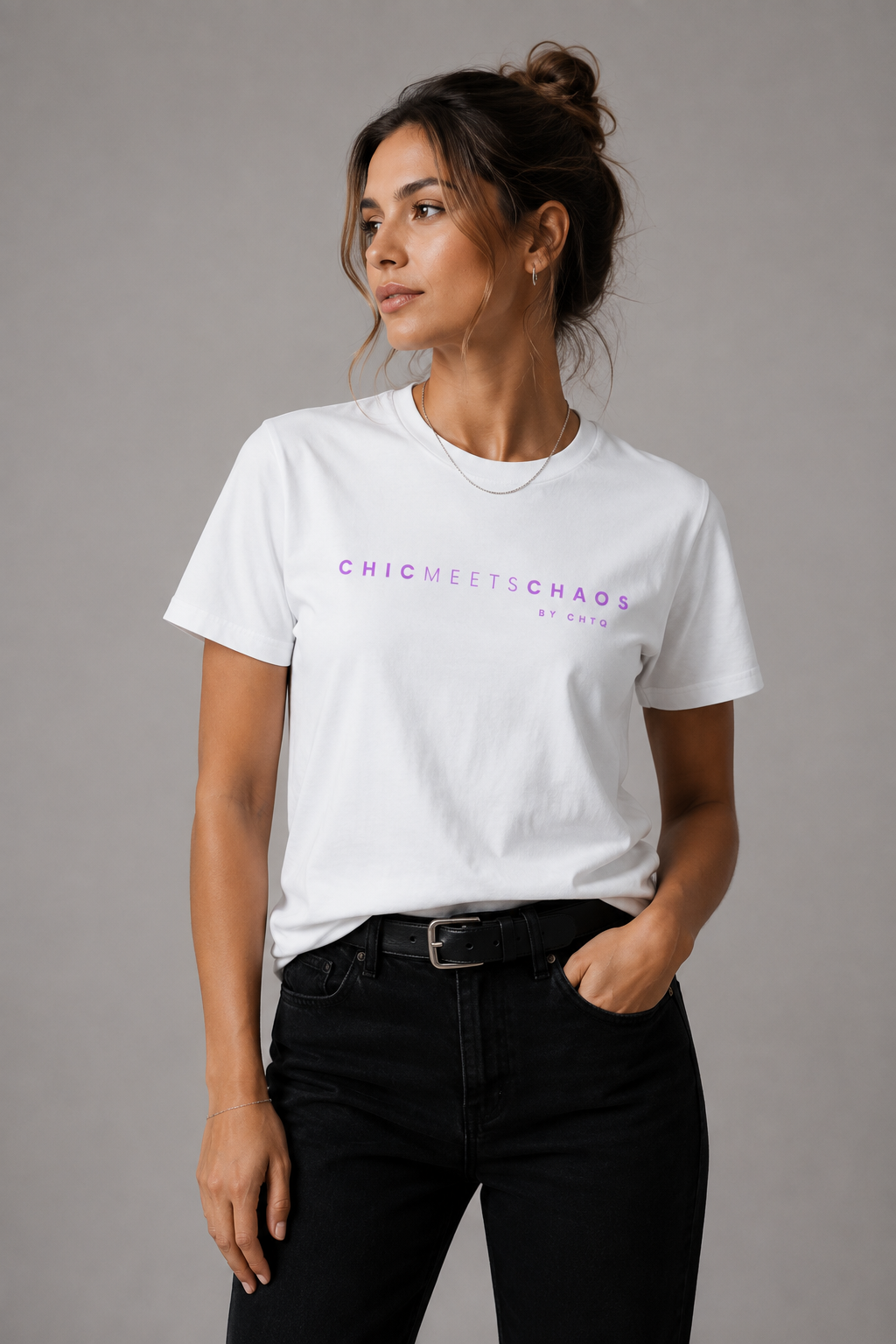 Woman wearing a white Chic Meets Chaos by CHTQ graphic T-shirt with purple typography, paired with black jeans against a gray studio background.