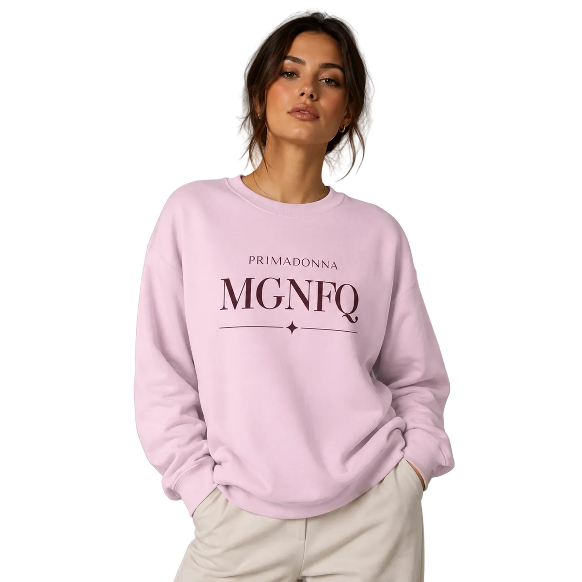 A woman wearing a light pink crewneck sweatshirt with 'PRIMADONNA MGNFQ' printed in dark pink on the front, paired with off-white trousers, against a solid gray studio background.
