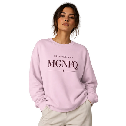 A woman wearing a light pink crewneck sweatshirt with 'PRIMADONNA MGNFQ' printed in dark pink on the front, paired with off-white trousers, against a solid gray studio background.