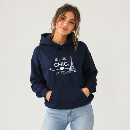 A woman wearing a navy blue hoodie with a white graphic print that reads 'JE SUIS CHIC. ET TOI?' with an Eiffel Tower illustration, paired with light blue jeans, against a plain light gray background.