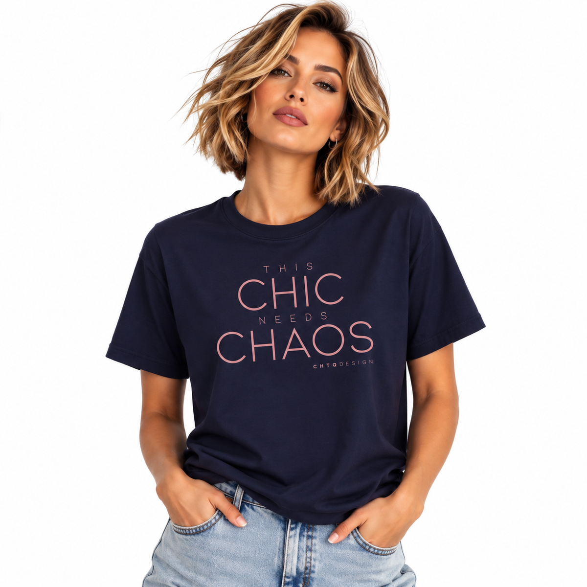 A woman wearing a navy blue graphic t-shirt with the text 'THIS CHIC NEEDS CHAOS' in pink, paired with light-wash blue jeans, against a plain white background.
