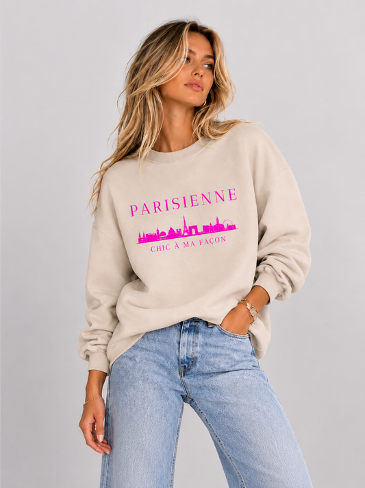 A woman wearing a cream crewneck sweatshirt with a bright pink “PARISIENNE” graphic, Paris skyline design, and “CHIC À MA FAÇON” text, paired with light-wash blue jeans against a neutral gray studio background.