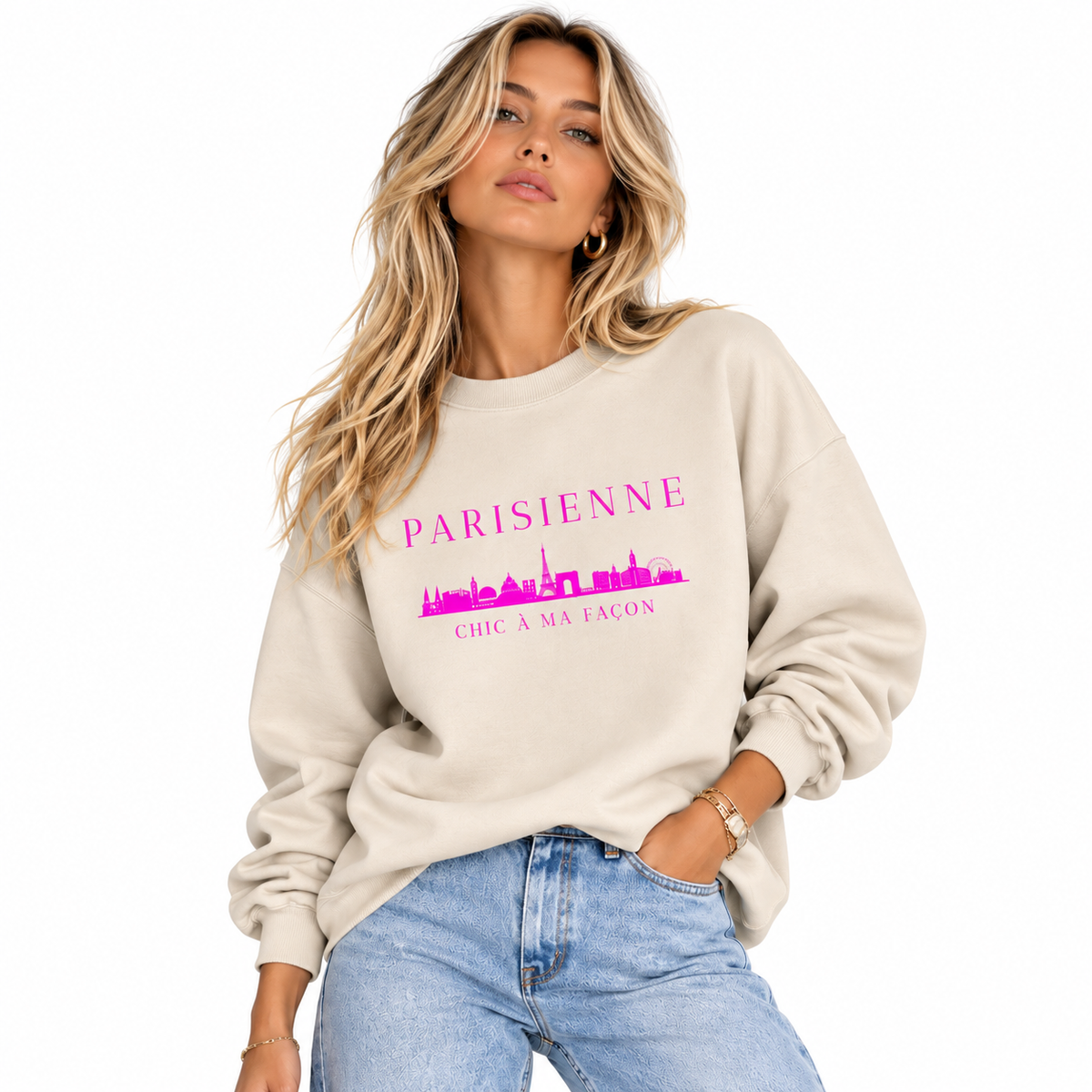 A woman wearing a beige crewneck sweatshirt with a pink graphic print that reads 'PARISIENNE' and 'CHIC A MA FACON' with a skyline illustration, paired with light blue jeans against a white background.
