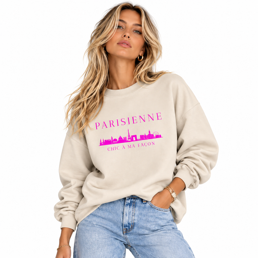 A woman wearing a beige crewneck sweatshirt with a pink graphic print that reads 'PARISIENNE' and 'CHIC A MA FACON' with a skyline illustration, paired with light blue jeans against a white background.