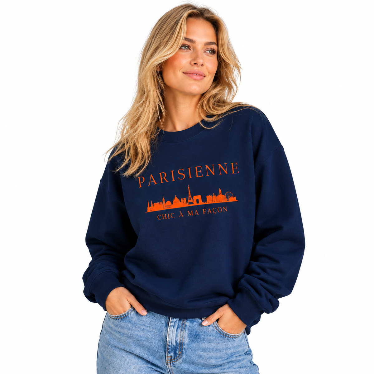 A woman wearing a navy blue crewneck sweatshirt with an orange graphic print that reads 'PARISIENNE CHIC A MA FACON' above an orange skyline illustration, paired with light blue jeans against a plain white background.