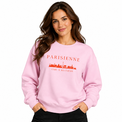 A woman modeling a light pink crewneck sweatshirt with a red graphic print that reads 'PARISIENNE' and 'CHIC A MA FACON' with a skyline illustration, set against a plain white background.