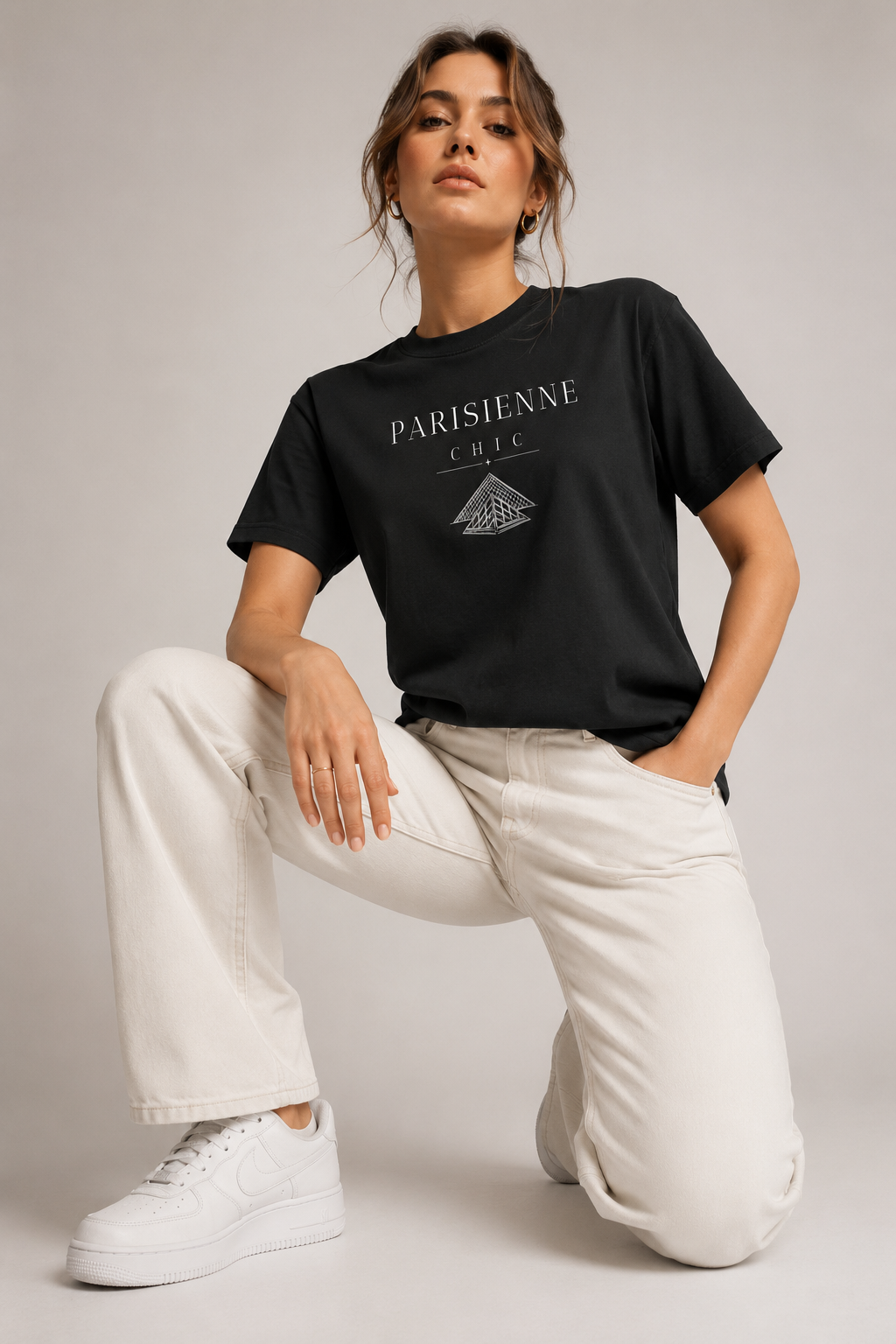 Woman wearing a black Parisienne Chic graphic T-shirt with a white Louvre Pyramid-inspired print, styled with cream trousers in a neutral studio setting.