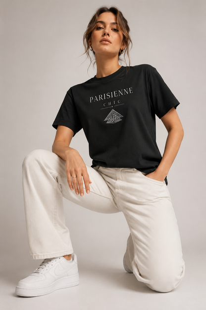 Woman wearing a black Parisienne Chic graphic T-shirt with a white Louvre Pyramid-inspired print, styled with cream trousers in a neutral studio setting.