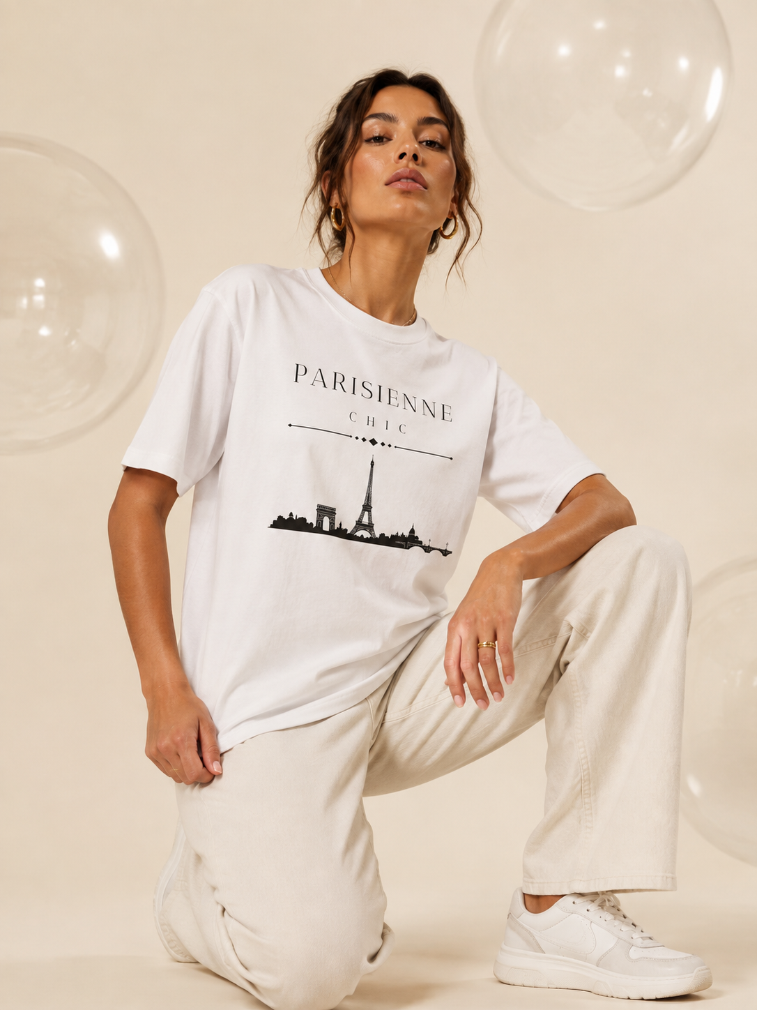A woman kneeling in a studio wearing an oversized white PARISIENNE CHIC graphic T-shirt with a black Paris skyline and Eiffel Tower print, paired with cream wide-leg trousers and white sneakers against a soft champagne background.