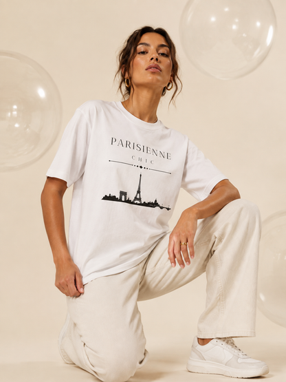 A woman kneeling in a studio wearing an oversized white PARISIENNE CHIC graphic T-shirt with a black Paris skyline and Eiffel Tower print, paired with cream wide-leg trousers and white sneakers against a soft champagne background.